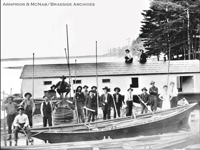 Pointer boat and crew of McLachlin Lumber Firm in Arnprior at the park, with McLachlin Mill visible in background. Photograph taken around 1901.