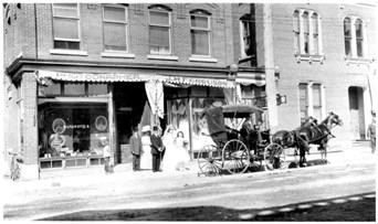 Photo of a wedding party in front of the Handford and J.M Ferguson stores.  Note on back of print says "Wedding Party (unknown) after having photos taken about 1905-06-07, Raglan St. Renfrew. Entrance to Albert Limmer's home - white brick building - part (present bowling alley)".