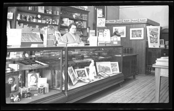 Thelma Stringer behind the sales desk of Handford Studio shop. Many frames and photographs visible for sale, as well as camera materials.