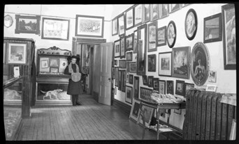 Lillian Handford in the shop with frames hanging on the walls, which were avaialble for sale. She is wearing a large brimmed hat and a fur cuff and scarf, leaning against a piece of furniture.